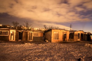 Nighttime scene of a row of shacks blanketed in snow, with soft moonlight casting shadows