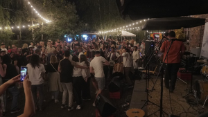 Na zewnątrz, w świetle lampionów, nocą tańczą ludzie, niektórzy grają na instrumentach. Na scenie stoją dwaj muzycy odwróceni tyłem.