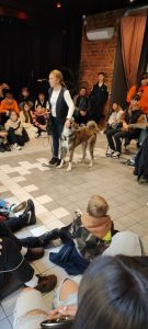 Grupowe zdjęcie ze stojącą na środku sali kobietą w blond włosach spiętych w kucyk, razem z psem.