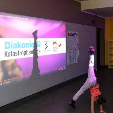 Dziewczynka w różowej bluzce i szarych spodniach stoi na rękach, po lewej widać fioletowy obraz z logotypami Diakonie Katastrophen, NOMADA oraz Diakonia, wyświetlony projektorem na ścianie