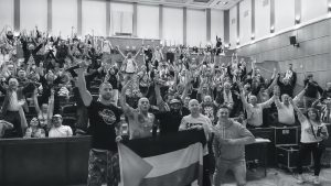 Widownia pełna ludzi z uniesionymi do góry rękami, na czele zdjęcia muzycy z flagą Palestyny.
