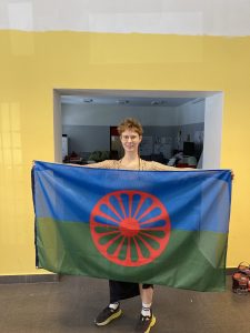 Na zdjęciu kobieta z krótkimi włosami i długim cienkim warkoczykiem trzyma romską flagę: niebiesko-zieloną z czerwonym kołem wozu na środku.