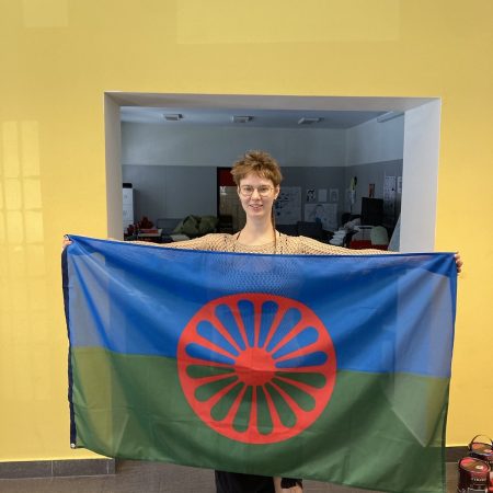 Na zdjęciu kobieta z krótkimi włosami i długim cienkim warkoczykiem trzyma romską flagę: niebiesko-zieloną z czerwonym kołem wozu na środku.
