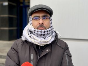 Mężczyzna w okularach szaliku i czapce, patrzący zamyślony w bok na jasnym tle.