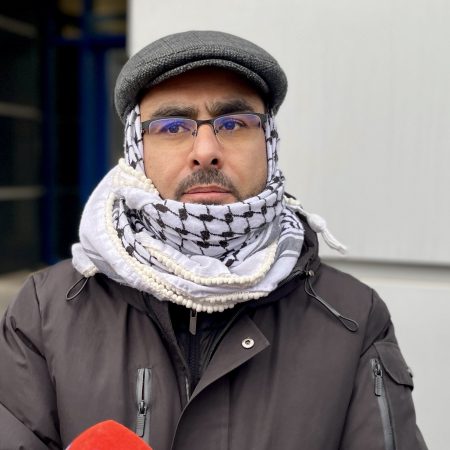 Mężczyzna w okularach szaliku i czapce, patrzący zamyślony w bok na jasnym tle.