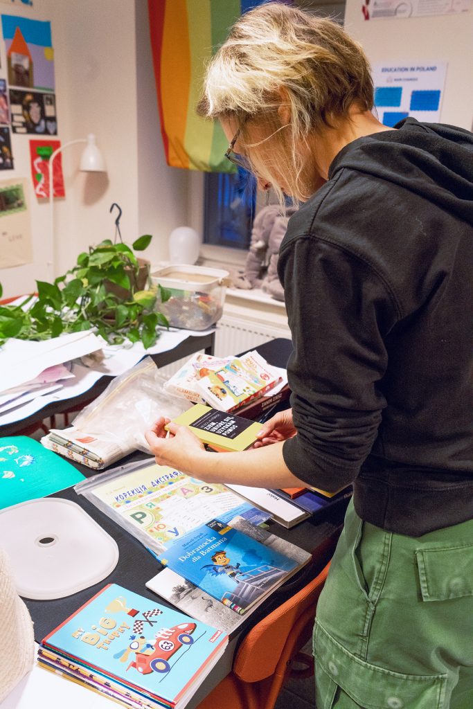 A woman focused on book at a desk in a bright room, surrounded by many books and educational materials.