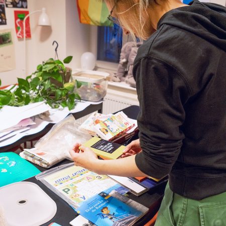 A woman focused on book at a desk in a bright room, surrounded by many books and educational materials.