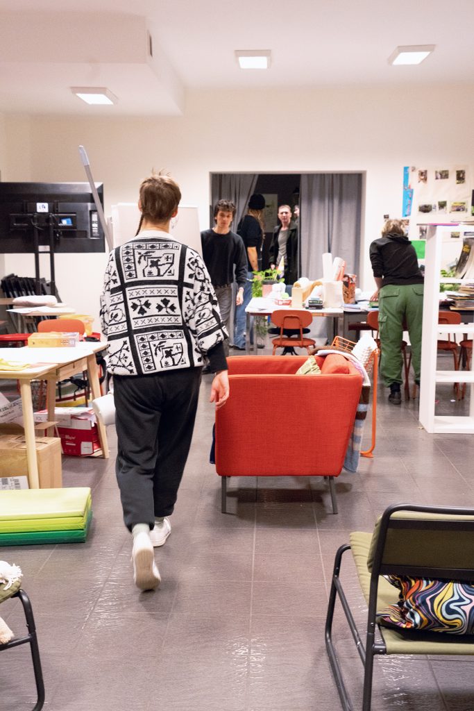 A woman walks through a room filled with furniture and people engaged in cleaning