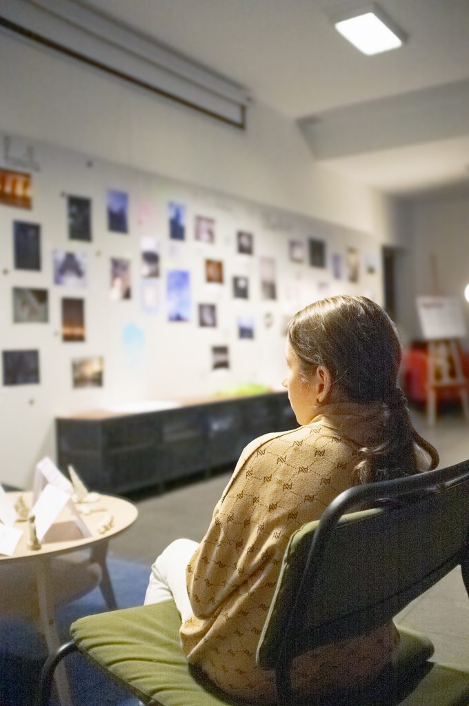 A girl with the ginger hair sits in a chair, gazing thoughtfully at framed pictures hanging on the wall.