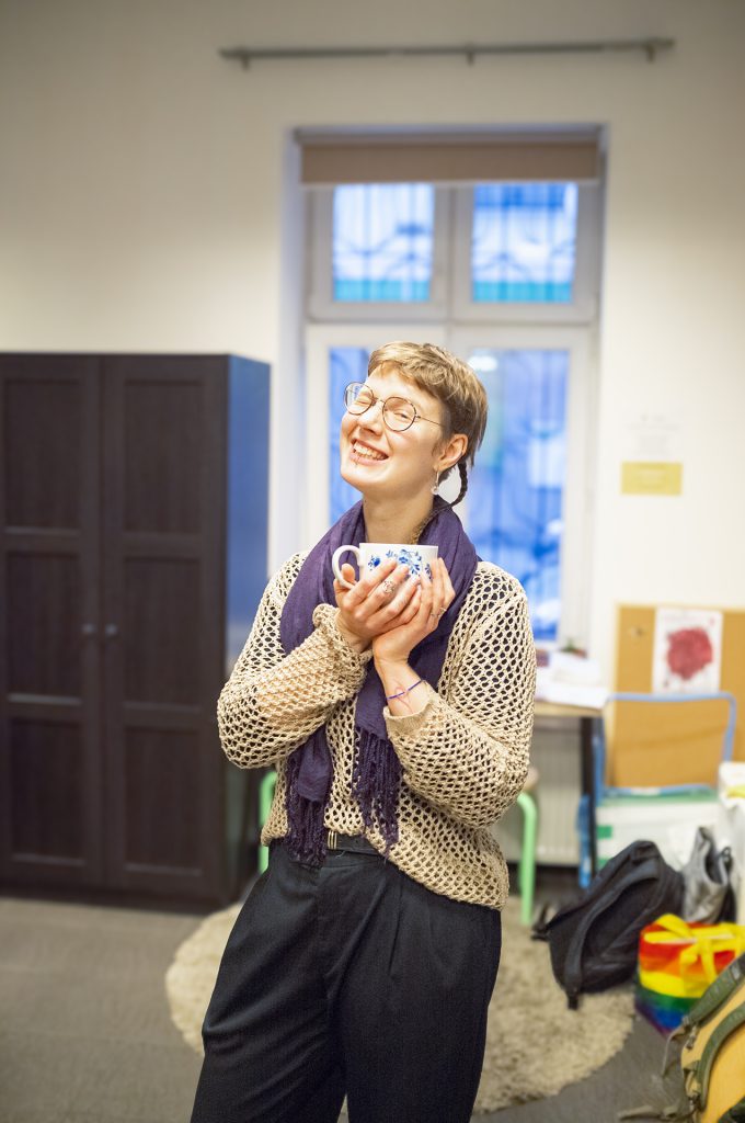 A smiling woman wearing glasses and a colorful scarf, holding a cup, radiating warmth and friendliness.