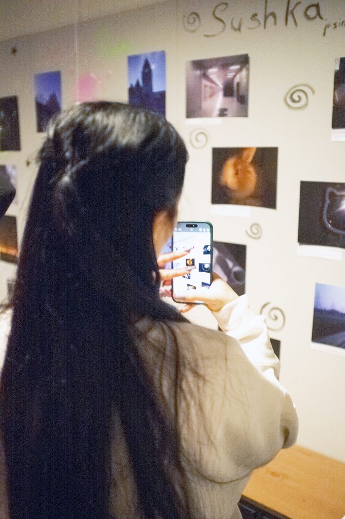 A woman photographing a wall adorned with various pictures, capturing the art displayed.
