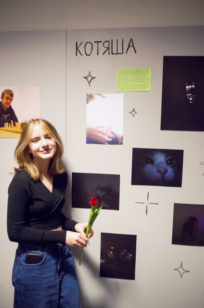 A teenager girl stands beside a wall adorned with various framed pictures, smiling as she admires her artwork.