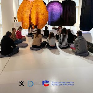 A group of teenagers sitting on the floor, focused on large orange and purple artistic objects, with the logos of Nomada, FED, and the Polish-American Freedom Foundation.
