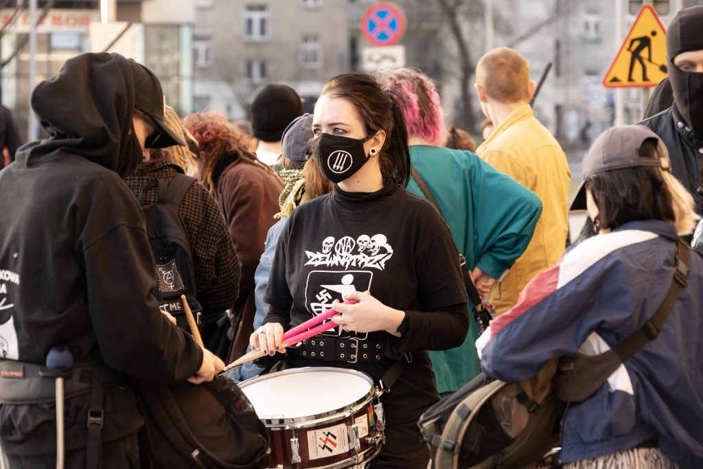 A woman in a mask i spreparing to play a drum on the protest, surrounded with people