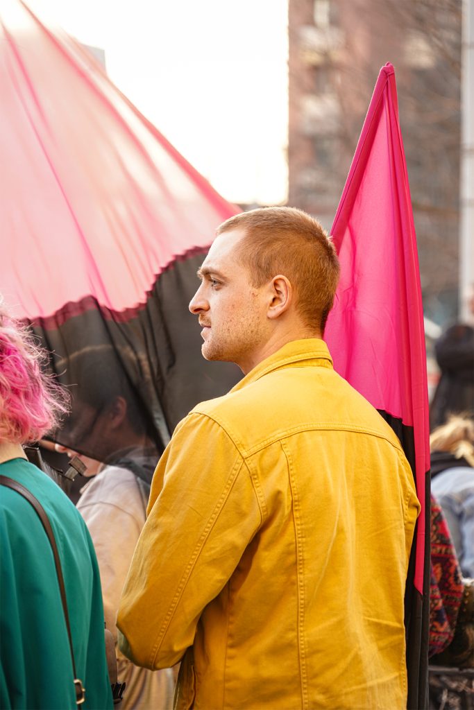 A man with short hair with vibrant red and black flag wearing a bright yellow jacket, smiling and standing in the sun surrounded with crowd