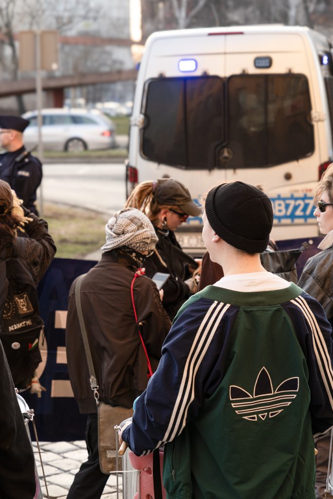 A woman with her back turned in a dark blue and green jacket with adidas logo stands confidently, police car in the background