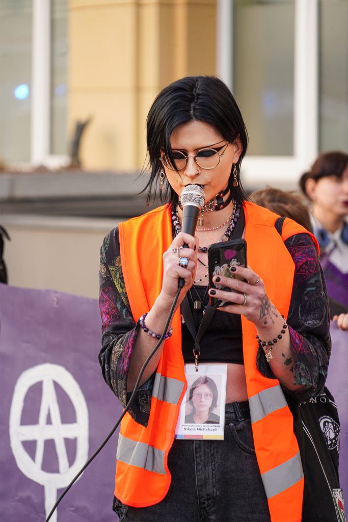 A woman wearing an orange vest stands confidently, holding a microphone as she speaks.