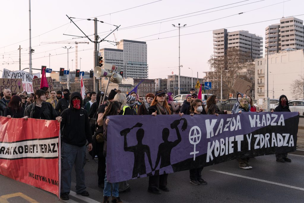 A diverse group of people stands on the street, proudly holding a large banner together with a sighn on it "Each authority cheats on women"