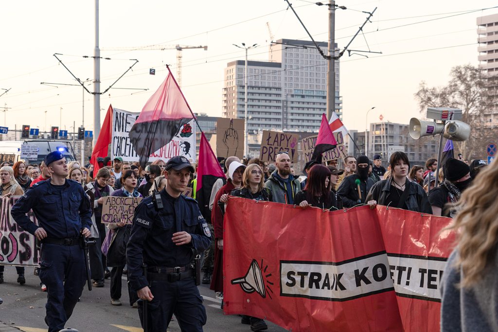 A diverse group of people walking together down the street, proudly holding a colorful banner "Woman's Strike", police in the foreground, walking along the street near the protesters
